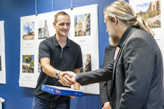 Mężczyzna w ciemnej marynarce wręcza nagrodę mężczyźnie w ciemnej koszulce. W tle plansze ze zdjęciami.