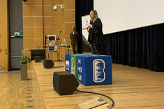 Na zdjęciu widzimy scenę w dużej sali audytoryjnej. Na środku sceny stoi kobieta ubrana na czarno, przemawiająca do publiczności. W tle widoczny jest mężczyzna w brązowej marynarce. Na scenie znajdują się także sprzęty techniczne – mikrofony, komputer oraz ekran projekcyjny, który zajmuje dużą część prawej strony kadru. Na podeście znajdują się niebieskie i zielone kostki z logotypami.