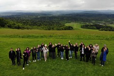 Zdjęcie wykonane z wysokości kilkudziesięciu metrów. Grupa około dwudziestu osób ubranych na sportowo pozuje do zdjęcia. Wszyscy spoglądają do góry i machają do obiektywu. W tle zielona polana i panorama gór.