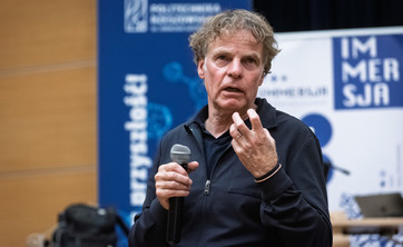 Mężczyzna, prelegent ubrany na czarno, podczas prowadzenia wykładu. W tle biało-niebieskie banery informacyjne. Prof. arch. Winy Maas, MVRDV