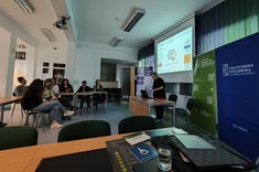 W lekko przyciemnionym pomieszczeniu widocznych jest po lewej stronie kilka osób słuchających wykładu. Po prawej stronie wysoko zawieszony jasny ekran, przed nim stoi prelegentka.