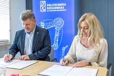 Od lewej rektor, prof. P. Koszelnik, dr. B. Szafrańska. Dwie osoby, kobieta i mężczyzna siedzą przy stole i podpisują umowę. Oboje elegancko ubrani, mężczyzna w ciemną marynarkę, kobieta na jasno. W tle niebieski baner z białym napisem Politechnika Rzeszowska.