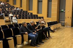 Zdjęcie ukazuje fragment audytorium podczas wydarzenia. Widzimy przestronną salę z nowoczesnym, drewnianym wystrojem ścian i dużymi, zasłoniętymi oknami. W centralnej części kadru siedzi grupa uczestników — w pierwszym rzędzie zasiada kilka osób. Są elegancko ubrani, a część z nich jest zaangażowana w rozmowę. 