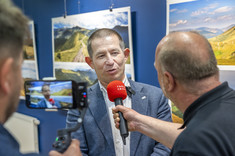 Mężczyzna w ciemnej marynarce udziela wywiadu dziennikarzowi z mikrofonem. Widoczne jest nagrywanie wywiadu na telefonie komórkowym. W tle na granatowej ścianie zawieszone plansze z dużymi kolorowymi fotografiami.