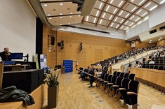 Zdjęcie przedstawia przestronną i nowoczesną salę audytoryjną podczas trwającego wydarzenia. Po lewej stronie, na scenie, stoi mężczyzna przemawiający do mikrofonu. Widownia jest częściowo zapełniona – uczestnicy siedzą w eleganckich, drewnianych fotelach z granatową tapicerką, a ich uwaga skupiona jest na prelegencie. Sala wyróżnia się nowoczesnym designem z drewnianymi panelami na ścianach i suficie, oraz starannie rozmieszczonym oświetleniem.