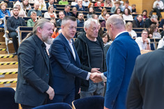 Czterech mężczyzn podczas oficjalnego przywitania. Trzech zwróconych przodem do zdjęcia. W tle uczestnicy wykładu. Od lewej: arch. W. Matłok, prof. J. Strojny, arch. W. Fałat, prez. K. Fijołek.