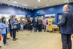 Ujęcie pomieszczenia, w którym w kilku grupach stoją uczestnicy wystawy. Wszyscy zwróceni w jednym kierunku, słuchają przemawiającego mężczyzny, widocznego po prawej stronie zdjęcia. W tle na granatowej ścianie zawieszone plansze z dużymi kolorowymi fotografiami.