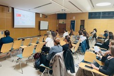 W jasnym wnętrzu sali wykładowej ze ścianami z drewnopodobną wykładziną i z białym sufitem zgromadzona kilkudziesięcioosobowa grupa słuchaczy. W tle jasny ekran z wyświetloną prezentacją. 