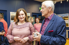 Na pierwszym planie w pomieszczeniu stoją dwie osoby, kobieta w różowej bluzce i mężczyzn w ciemnej marynarce. Rozmawiają ze sobą. W tle stoi kilka osób.