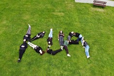 Zdjęcie wykonane z wysokości kilku metrów. Na zielonej trawie leży kilka osób , których sylwetki tworzą napis KUA.