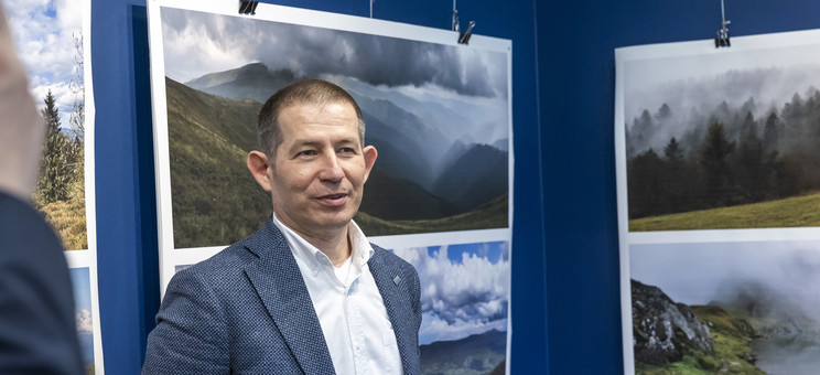 Na pierwszym planie stoi autor wystawy, mężczyzna w ciemnej marynarce.  W tle na granatowej ścianie zawieszone plansze z dużymi kolorowymi fotografiami pejzaży górskich.