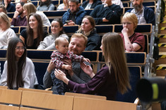 Zbliżenie na uczestników wykładu siedzących w rzędach. Pośrodku mała dziewczynka, najmłodszy uczestnik wykładu. Wszyscy się uśmiechają. Na pierwszym planie studenci kierunku architektura.