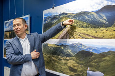 Na pierwszym planie stoi autor wystawy, mężczyzna w ciemnej marynarce, wskazuje ręką na fotografię. W tle na granatowej ścianie zawieszone plansze z dużymi kolorowymi fotografiami pejzaży górskich.