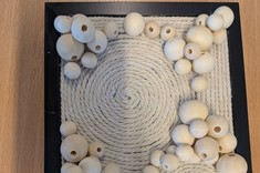 Na jasnym stole leży praca plastyczna. Na czarnej kwadratowej podstawie jasnym sznurkiem ukształtowane koła, linie, pomiędzy nimi linie z różnej wielkości koralików drewnianych.