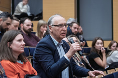 Zbliżenie na słuchaczy wykładu siedzących w rzędach. Po środku uczestnik wykładu, mężczyzna w ciemnej marynarce podczas zadawania pytania prelegentowi.