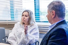 Mężczyzna i kobieta siedzą przy stole podczas rozmowy. Mężczyzna w ciemnej marynarce, kobieta w białej bluzce. W tle przeszklona ściana.