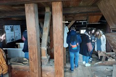 Zdjęcie przedstawia kameralne wnętrze w drewnianym budynku. Widoczne na pierwszym planie drewniane słupy. Wnętrze wypełnione sprzętami używanymi dawniej w gospodarstwie. Grupa kilku studentów ogląda wystawione eksponaty.