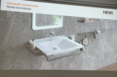 Na zdjęciu ekran z wyświetloną prezentacją. Na slajdzie widoczne wnętrze łazienki. Na tle szarej ściany biała umywalka z bocznymi uchwytami.