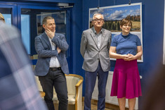 W pomieszczeniu stoją obok siebie trzy osoby, dwóch mężczyzn i kobieta po lewej stronie. Wszyscy zwróceni w jednym kierunku. W tle na granatowej ścianie zawieszone plansze z dużymi kolorowymi fotografiami.