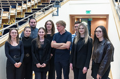 Zdjęcie grupowe grupy dziewięciu osób, kobiet i mężczyzn. Większość ubrana na czarno. W tle wyjście z sali wykładowej. Pośrodku prof. arch. W. Maas oraz jeden z opiekunów koła arch. M. Bober i członkowie koła naukowego IMMERSJA.