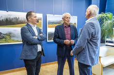 Trzech mężczyzn, ubranych w różnych odcieniach niebieskiego garniturach stoi obok siebie i rozmawia. Uśmiechają się do siebie. W tle na granatowej ścianie zawieszone plansze z dużymi kolorowymi fotografiami.