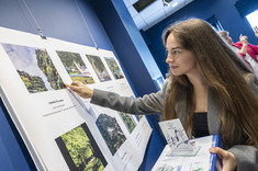 Zbliżenie na młoda kobietę z długimi włosami. Kobieta wskazuje ręką na zdjęcie, w drugiej ręce trzyma nagrodę. Na zdjęciu: stud. arch. I. Gruszka.