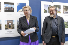 Kobieta i mężczyzna w ciemnych marynarkach stoją na tle plansz ze zdjęciami. Kobieta trzyma w ręku białą kopertę. Na zdjęciu: prof. M. Kłos, arch. W. Matłok.