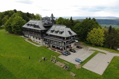 Zdjęcie wykonane z dużej wysokości. Na zdjęciu kilkukondygnacyjny budynek ze spadzistym dachem.  W tle ściana wysokich drzew, a w oddaleniu zarys gór.