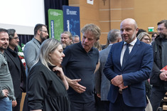 Kilka grup osób zgromadzonych podczas swobodnych rozmów w sali wykładowej. Na pierwszym planie grupa trzech osób, od lewej strony kobieta i mężczyzna ubrani na czarno i mężczyzna w ciemnym garniturze. Na pierwszym planie od lewej: prof. arch. A. Sikora, prof. arch. W. Maas, prez. K. Fijołek.