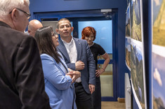 Kilka osób zgromadzonych przy planszach z fotografiami zawieszonymi na ciemnej ścianie. Przyglądają się uważnie szczegółom zdjęć.