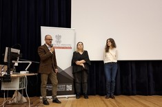 Zdjęcie przedstawia trzy osoby stojące na scenie podczas wydarzenia. Po lewej stronie, znajduje się mężczyzna w brązowej marynarce, który mówi do mikrofonu. Obok niego stoją dwie kobiety – jedna w czarnej bluzce i ciemnych spodniach, druga w jasnej koszuli i jeansach.
