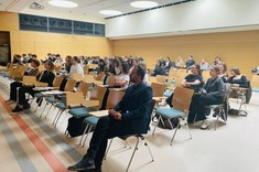 W jasnym wnętrzu sali wykładowej ze ścianami z drewnopodobną wykładziną i z białym sufitem zgromadzona kilkudziesięcioosobowa grupa słuchaczy. Wszyscy zajmują miejsca siedzące.