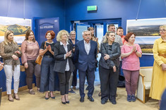 Ujęcie pomieszczenia w którym stoi grupa kilkunastu osób. Wszyscy zwróceni w jednym kierunku, biją brawo i uśmiechają się. W tle na granatowej ścianie zawieszone plansze z dużymi kolorowymi fotografiami.
