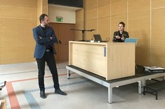 W jasnym wnętrzu sali wykładowej ze ścianami z drewnopodobną wykładziną na podwyższeniu za stołem siedzi prelegentka prowadząca wykład, przed stołem stoi mężczyzna słuchający wykładu. 