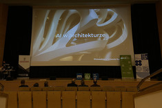 Na zdjęciu widzimy scenę w nowoczesnej sali audytoryjnej. Na dużym ekranie wyświetlony jest tytuł prezentacji. Tłem slajdu jest futurystyczne zdjęcie wnętrza budynku o nowoczesnych, organicznych kształtach.