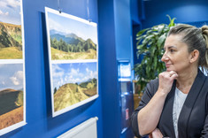 Przed planszami z kolorowymi fotografiami pejzaży górskich stoi kobieta w ciemnej marynarce. Przygląda się wystawie i uśmiecha się.