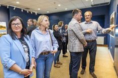 W pomieszczeniu na pierwszym planie stoją dwie kobiety. Obie ubrane na niebiesko. Jedna uśmiecha się do zdjęcia. W tle dwóch mężczyzn stoi przed prezentowanymi planszami i wskazują na zdjęcia.