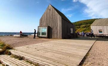 Zdjęcie: Pośrodku rozległej przestrzeni w naturalnym surowym krajobrazie zlokalizowany parterowy budynek. Bryła prosta z wysokim dachem, całość wykończona pionowym szarym deskowaniem. W tle zielone wzgórze i jaskrawo niebieskie niebo.