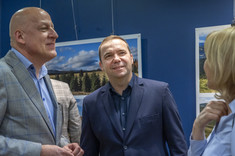 Trzy osoby rozmawiają ze sobą, dwóch mężczyzn i kobieta. Uśmiechają się. W tle na granatowej ścianie zawieszone plansze z dużymi kolorowymi fotografiami.