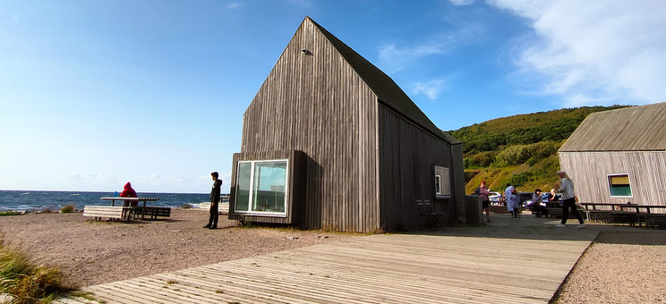 Zdjęcie: Pośrodku rozległej przestrzeni w naturalnym surowym krajobrazie zlokalizowany parterowy budynek. Bryła prosta z wysokim dachem, całość wykończona pionowym szarym deskowaniem. W tle zielone wzgórze i jaskrawo niebieskie niebo.