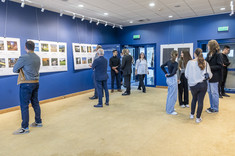Wnętrze z ciemno niebieskimi ścianami. Na ścianach zawieszone białe plansze z kolorowymi zdjęciami. We wnętrzu, w kilku luźnych grupach, zgromadzone osoby odwiedzjące wystawę.