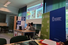 W lekko przyciemnionym pomieszczeniu po środku wysoko zawieszony jasny ekran, przed nim przy stole siedzą dwie prelegentki. W tle kilka wysokich na około 2 metry plansz w zielonym lub niebieskim kolorze z białymi napisami.