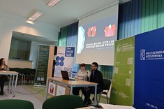 W lekko przyciemnionym pomieszczeniu po środku wysoko zawieszony jasny ekran, przed nim przy stole siedzi dwóch prelegentów, mężczyzna i kobieta. W tle kilka wysokich na około 2 metry plansz w zielonym lub niebieskim kolorze z białymi napisami.