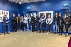 Zdjęcie uczestników wystawy. Grupa kilkunastu osób zgromadzona w pomieszczeniu na tle ciemno niebieskiej ściany.