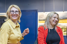 Dwie kobiety stoją obok siebie i uśmiechają się do fotografii. Obie z jasnym włosami, jedna w żółtej sukience, druga w czerwonej marynarce.