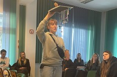 W pomieszczeniu na pierwszym planie stoi kobieta, w wysoko uniesionej ręce trzyma pracę plastyczną i tłumaczy coś zebranym za nią słuchaczom.