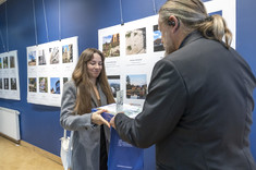 Mężczyzna ustawiony plecami do zdjęcia wręcza nagrodę młodej kobiecie z długimi włosami. W tle plansze ze zdjęciami. Na zdjęciu: stud. arch. I. Gruszka i arch. W. Matłok.