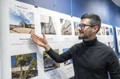 Zbliżenie na mężczyznę w czarnym golfie i w okularach. Mężczyzna wskazuje ręką na zdjęcie. Na zdjęciu: arch. J. Dudek.