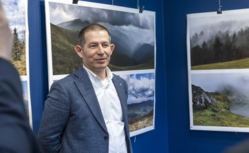 Na pierwszym planie stoi autor wystawy, mężczyzna w ciemnej marynarce.  W tle na granatowej ścianie zawieszone plansze z dużymi kolorowymi fotografiami pejzaży górskich.