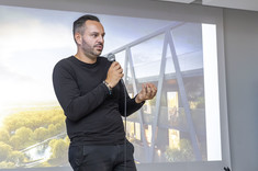 Prelegent Maciej Franta opowida o swoim projekcie. Na tle jego postaci znajduje się slajd z wizualizacją budynku.